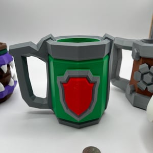 May include: A green, low-poly mug featuring a grey handle and rim, with a red shield emblem on the front. A silver-colored US quarter coin is placed in front of the mug to provide a sense of scale for the object's physical dimensions.