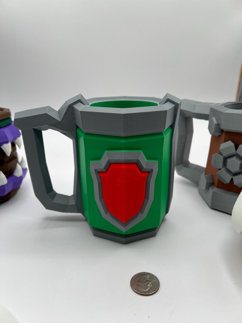 May include: A green, low-poly mug featuring a grey handle and rim, with a red shield emblem on the front. A silver-colored US quarter coin is placed in front of the mug to provide a sense of scale for the object's physical dimensions.