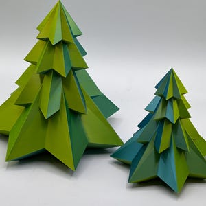 Puede incluir: Dos árboles de Navidad decorativos y geométricos en tonos verdes y turquesas. El árbol más grande es principalmente verde, mientras que el más pequeño tiene detalles turquesas. Ambos árboles tienen un diseño angular y en capas.