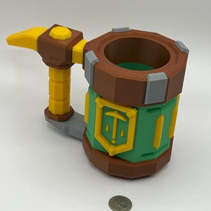 May include: A multi-colored 3D printed mug featuring a brown rim and base, a green cylindrical body with a yellow shield emblem displaying a pickaxe icon, and a yellow handle shaped like a hammer. A US quarter coin is placed in front for scale.
