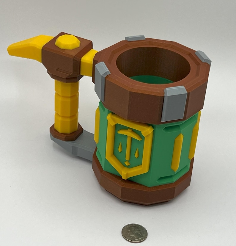 May include: A multi-colored 3D printed mug featuring a brown rim and base, a green cylindrical body with a yellow shield emblem displaying a pickaxe icon, and a yellow handle shaped like a hammer. A US quarter coin is placed in front for scale.