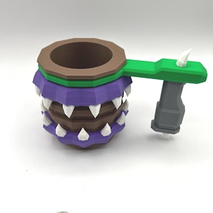 May include: A brown and purple mug with a faceted design, featuring white triangular teeth around the exterior. It has a green handle attached to a grey vertical grip. A silver US quarter coin is placed nearby for scale, measuring 0.955 inches.