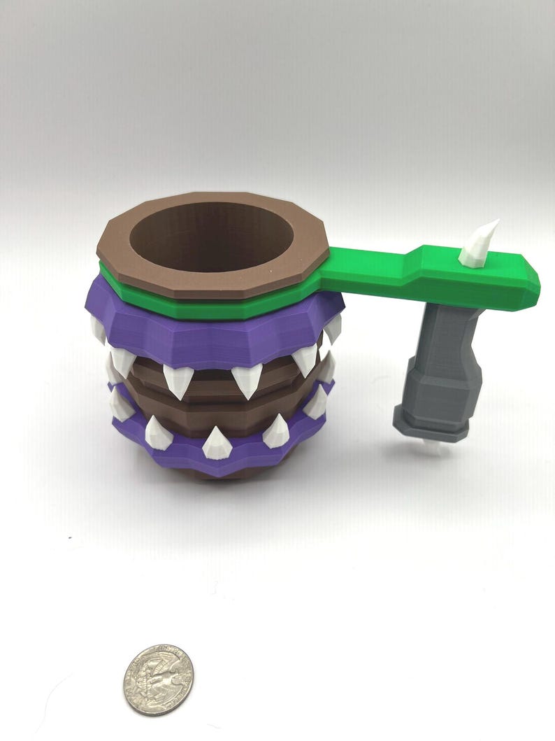 May include: A brown and purple mug with a faceted design, featuring white triangular teeth around the exterior. It has a green handle attached to a grey vertical grip. A silver US quarter coin is placed nearby for scale, measuring 0.955 inches.