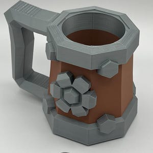May include: A brown and gray mug with a hexagonal shape, featuring a large gray handle and gray geometric accents resembling bolts and a gear on the exterior. A silver-colored US quarter coin is placed in front for scale reference.