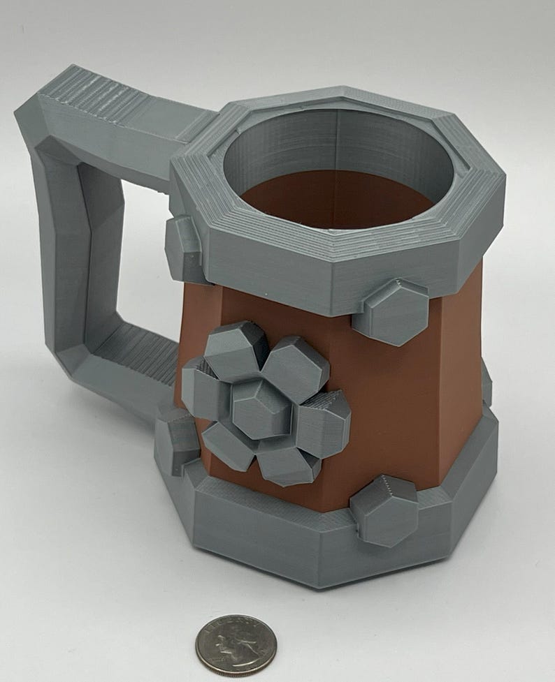 May include: A brown and gray mug with a hexagonal shape, featuring a large gray handle and gray geometric accents resembling bolts and a gear on the exterior. A silver-colored US quarter coin is placed in front for scale reference.
