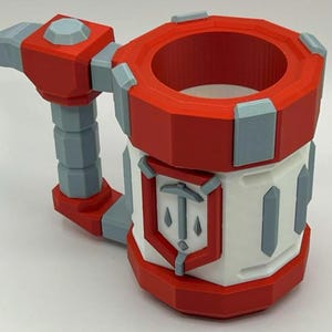May include: A cylindrical mug featuring a red and white color scheme with light gray accents. The mug has a thick handle and a prominent shield-shaped emblem on the front displaying a pickaxe icon. The design has a geometric, faceted appearance.