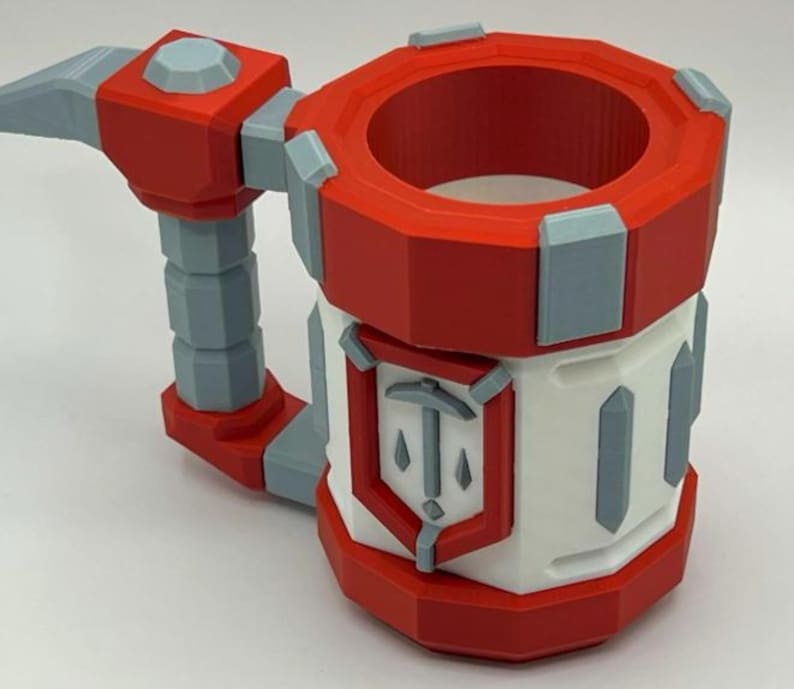 May include: A cylindrical mug featuring a red and white color scheme with light gray accents. The mug has a thick handle and a prominent shield-shaped emblem on the front displaying a pickaxe icon. The design has a geometric, faceted appearance.