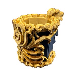 May include: A cylindrical cup featuring a dark blue interior and exterior base, adorned with intricate, three-dimensional gold-colored filigree overlays. The decorative gold elements include a prominent skull emblem on the side and ornate, swirling patterns that wrap around the upper rim and body of the vessel.