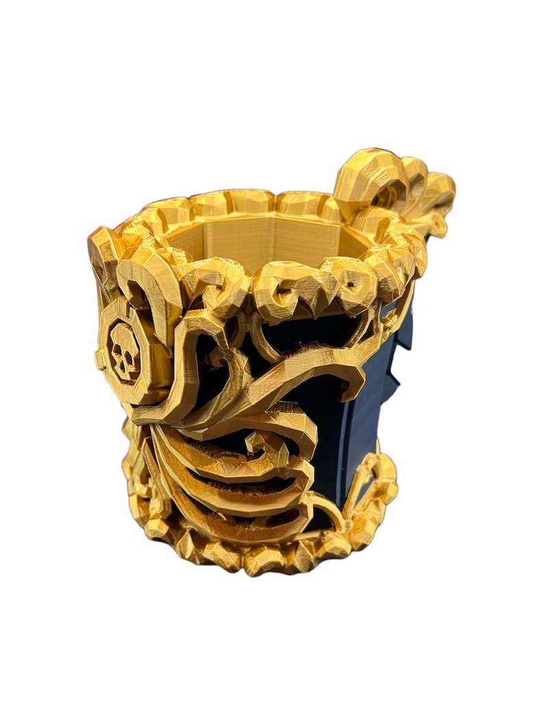 May include: A cylindrical cup featuring a dark blue interior and exterior base, adorned with intricate, three-dimensional gold-colored filigree overlays. The decorative gold elements include a prominent skull emblem on the side and ornate, swirling patterns that wrap around the upper rim and body of the vessel.