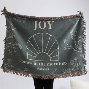 May include: A green woven throw blanket with a white sun design and the text "JOY comes in the morning: Psalm 30:5".