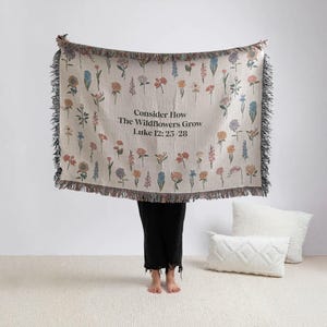 May include: A beige throw blanket with a floral pattern and black fringe. The blanket features various colorful wildflowers and the text "Consider How The Wildflowers Grow Luke 12: 25-28".