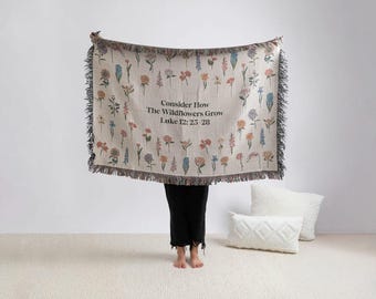 May include: A beige throw blanket with a floral design and black fringe. The blanket displays a variety of colourful wildflowers and the text "Consider How The Wildflowers Grow Luke 12: 25-28".