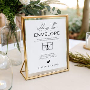 May include: A white rectangular sign in a gold metal frame featuring black text that reads address The ENVELOPE, PLEASE HELP THE BUSY-TO-BE BY MAKING YOURSELF THE ADDRESSEE, THANK YOU FOR CELEBRATING OUR SPECIAL DAY WITH US!, and OLIVER & AMELIA.