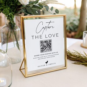 May include: A gold-framed sign with white paper featuring the black text Capture THE LOVE, a QR code, and the names OLIVER & AMELIA. The sign includes instructions to share photos by scanning the code with a smartphone and uploading images.