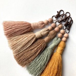May include: A set of four tassel keychains with wooden beads and copper-toned metal clasps. The tassels are in shades of pink, beige, green, and yellow.