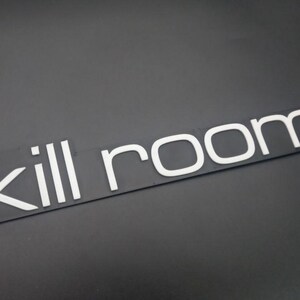 Op de afbeelding: Een rechthoekig zwart bord met de witte tekst "kill room" in een strak, schreefloos lettertype. De letters zijn verhoogd en duidelijk leesbaar tegen de donkere achtergrond, wat zorgt voor een visueel display met hoog contrast.