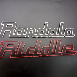 May include: Two stylized name decals. The top decal reads "Randala" in white with a black outline. The bottom decal reads "Riddle" in red with a black outline. Both decals are in a cursive font and are set against a dark gray background.