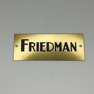 May include: A rectangular brass nameplate with the name "FRIEDMAN" in black, centered. The plate has two small holes, one on each end. The background is a light gray.
