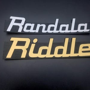 May include: Two black rectangular signs with the words "Randala" in white and "Riddle" in gold.