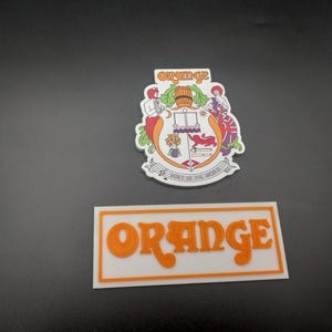 Op de afbeelding: Twee witte rechthoekige borden met oranje tekst. Het bovenste bord heeft een wapenschild met een vat, een kroon en de tekst "Orange" boven het wapenschild. Het onderste bord heeft de tekst "Orange" in een gestileerd lettertype.