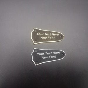 Puede incluir: Dos placas de clavijero de guitarra con el texto "Your Text Here Any Font". Una placa es de color gris oscuro con texto blanco y la otra es negra con texto blanco. Ambas tienen un borde blanco y tres agujeros.