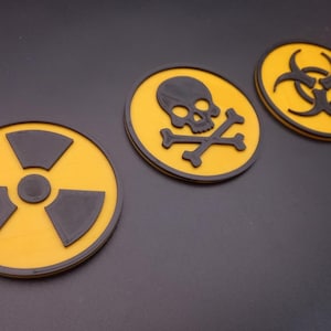 Könnte beinhalten: Drei gelbe und schwarze Warnschilder. Die Schilder sind rund und zeigen ein Strahlungssymbol, einen Totenkopf mit Knochen und ein Biohazard-Symbol.