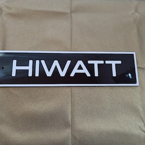 Może przedstawiać: Czarny prostokątny znak z białym napisem "HIWATT".