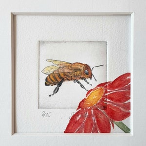 May include: A framed watercolor print of a bee hovering near a red flower. The bee is detailed with brown and yellow markings. The flower has red petals and a yellow center. The print is numbered 8/35.