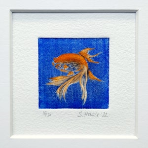 May include: Framed artwork featuring a detailed illustration of a goldfish. The fish is orange and white, set against a blue background. The artwork is signed and numbered 18/50. The frame is white.