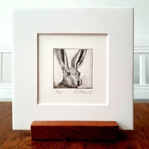 Könnte beinhalten: Eine Schwarzweiß-Radierung eines Hasenkopfes, gerahmt in Weiß mit einem Holzständer. Die Radierung ist signiert mit "S.House '22" und nummeriert "5/65".