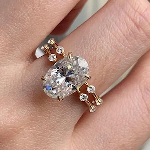 May include: A gold ring featuring an oval-cut diamond as the primary stone. The ring has a delicate band with smaller, round diamonds set along the sides. The ring is shown on a person's finger.