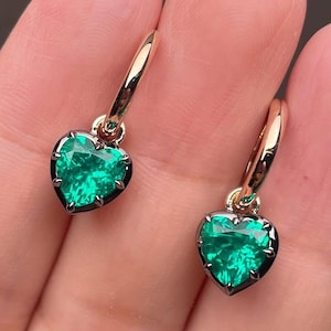 May include: A pair of gold hoop earrings, each with a heart-shaped, vivid green gemstone. The gemstones are set in a dark metal, contrasting with the gold hoops. The earrings are shown against a neutral background.