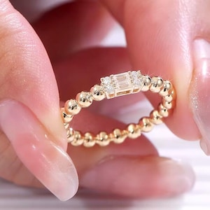 Könnte beinhalten: Ein goldener Ring mit einem perlenbesetzten Band und einem rechteckigen Mittelstein, der von kleineren runden Steinen flankiert wird. Der Ring wird von einer Hand mit gepflegten Nägeln gehalten. Der Ring ist ein Schmuckstück.