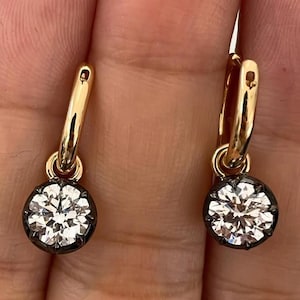 May include: A pair of gold hoop earrings featuring a round, white gemstone pendant. The gemstone is set in a dark metal bezel. The earrings are shown against a neutral background.