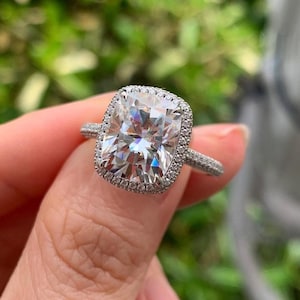 May include: A sparkling, square-cut diamond ring with a halo of smaller stones. The ring features a silver band with delicate detailing. The large center stone is the focal point, reflecting light and creating a brilliant display. The ring is held up by a person's hand.