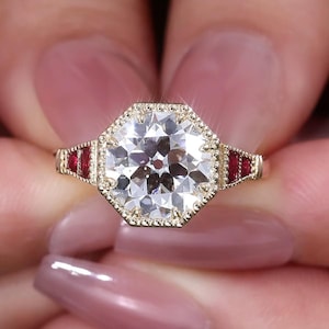 May include: A gold ring with a large, clear, octagonal-cut gemstone as the centerpiece. The ring features a halo of small, round stones around the main gem and red stones on each side of the band. The ring is held between two fingers.