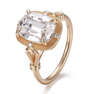 May include: A gold-colored ring with a large, clear, square-cut gemstone. The ring has a detailed, textured setting with small accent stones. The band is slender and polished, with the marking "18K" visible.