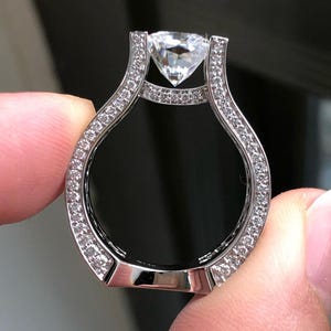 May include: A silver ring featuring a large, clear, diamond-shaped central stone. The band is set with numerous small, clear stones. The ring is held between fingers against a dark backdrop, highlighting the design and sparkle of the ring.