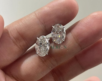 May include: A pair of oval-shaped diamond earrings set in a silver metal setting. The earrings are on a hand.