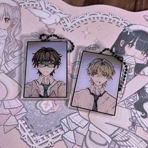 Puede incluir: Dos llaveros rectangulares con ilustraciones de estilo anime de personajes masculinos. Uno tiene cabello oscuro y gafas, el otro cabello claro. Ambos tienen cadenas de cuentas negras. Sobre una superficie rosa estampada.
