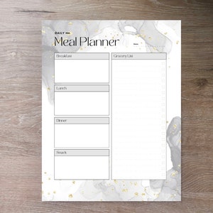May include: A white meal planner with a gray and gold marble design. The planner has sections for breakfast, lunch, dinner, and snack. It also includes a grocery list with checkboxes.