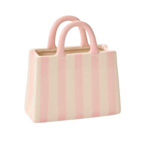 May include: A ceramic handbag-shaped vase with a pink and cream striped design. The vase features two pink handles and a rectangular form. The design is simple and elegant, suitable for home decor.