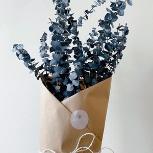 K&ouml;nnte beinhalten: Ein Strau&szlig; blauer Eukalyptus-Zweige, verpackt in braunes Papier mit einer wei&szlig;en Kordelschleife und einem Aufkleber mit der Aufschrift "Estrella Flowers & Gifts".
