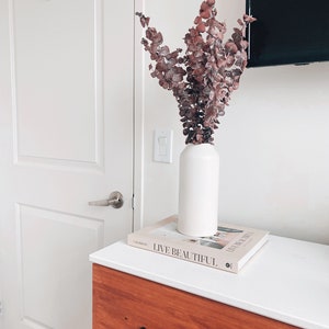 K&ouml;nnte beinhalten: Eine wei&szlig;e Keramikvase mit getrockneten Eukalyptus Zweigen in Rosa- und Braunt&ouml;nen, die auf einem Buch mit dem Titel "Live Beautiful" auf einer Kommode aus Holz mit wei&szlig;er Oberfl&auml;che steht.