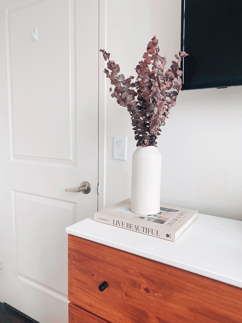 K&ouml;nnte beinhalten: Eine wei&szlig;e Keramikvase mit getrockneten Eukalyptus Zweigen in Rosa- und Braunt&ouml;nen, die auf einem Buch mit dem Titel "Live Beautiful" auf einer Kommode aus Holz mit wei&szlig;er Oberfl&auml;che steht.