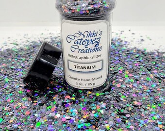 May include: A clear jar of chunky, hand-mixed holographic glitter labeled "Titanium" by Nikki's Pateyez Creations. The glitter is a mix of silver, black, and iridescent colors. The jar is open, with the lid beside it, and the glitter is scattered around.