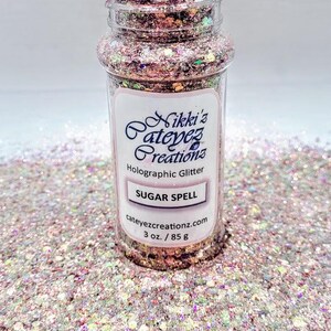 Może przedstawiać: Przezroczysty plastikowy słoik wypełniony holograficznym brokatem z etykietą "Sugar Spell" firmy Nikki's Cateyez Creations. Brokat to mieszanka różowych, srebrnych i opalizujących kolorów. Słoik zawiera 85 g brokatu.