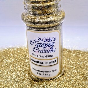 Puede incluir: Un frasco de vidrio transparente lleno de purpurina dorada, etiquetado como "Chandelier Mist" y "Ultra Fine Glitter". El frasco contiene 85 g de purpurina. La purpurina está esparcida alrededor del frasco, creando un efecto brillante.