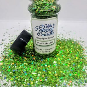Peut inclure: Un pot en plastique transparent rempli de paillettes holographiques vertes et irisées. Le pot a un couvercle noir et une étiquette qui indique "Nikki's atepez Creations Holographic Glitter SPLASH OF LIME Chunky Hand-Mixed 3 oz./85 g".