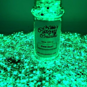 May include: A clear plastic jar filled with glowing green glitter, surrounded by a pile of the same luminous material. The label reads Nikki's Cateyez Creations, Glow Glitter, PIXIE DUST, and cateyezcreationz.com in black text on a white background.